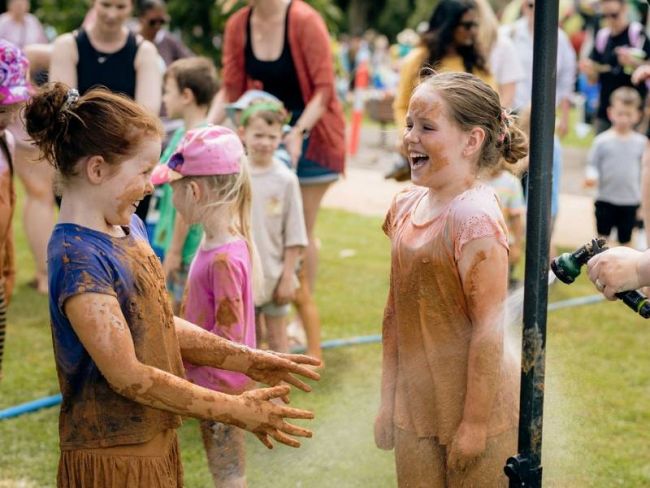 Two young children covered in mud being sprayed with water