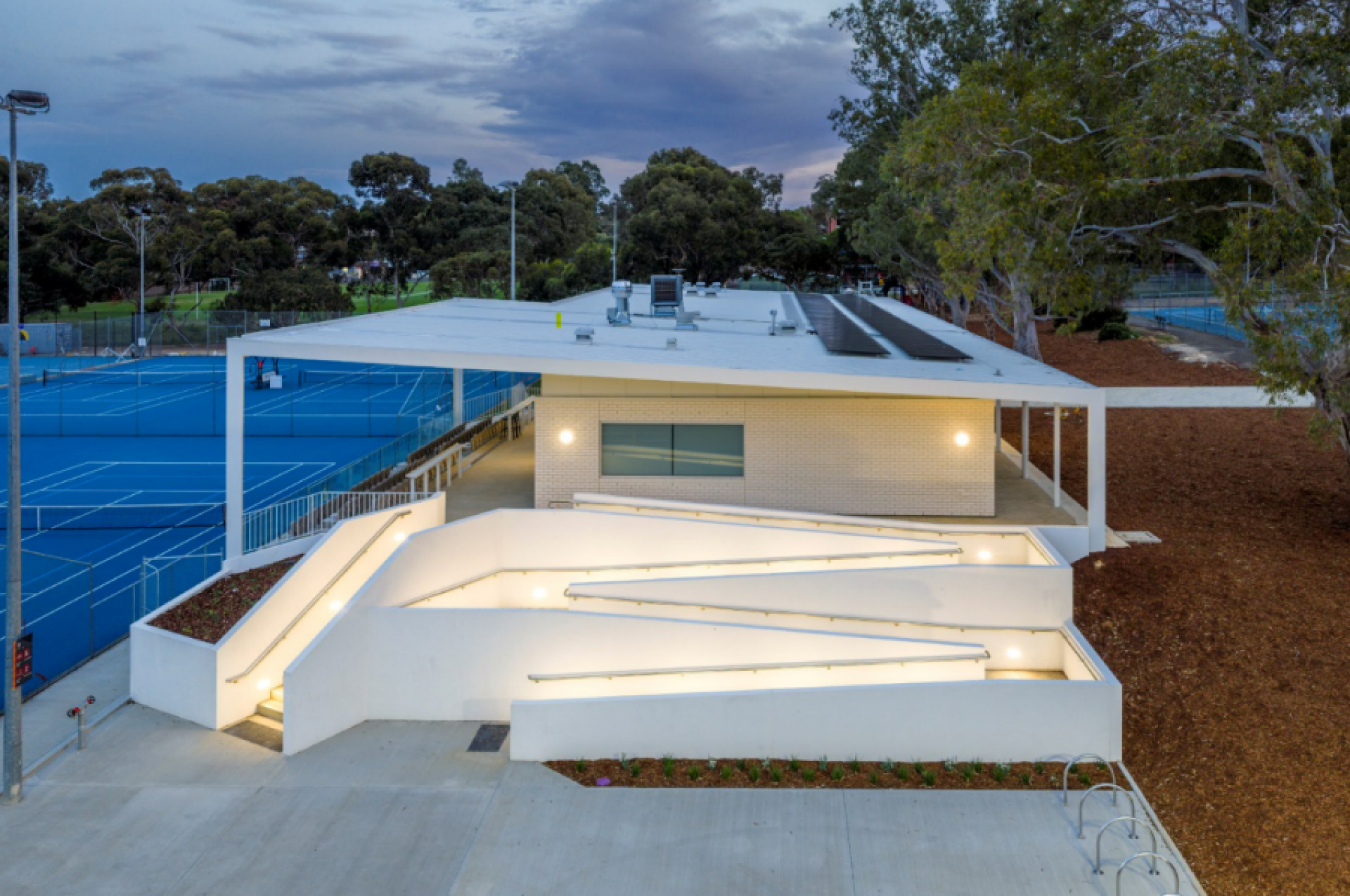 Tennis club building with illuminated zigzag accessibility ramp, adjacent to tennis courts